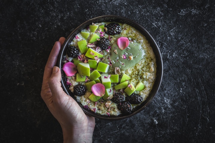 Hand howling bowl of cardamom rose porridge with matcha dust, sprouted pumpkin seed butter, green apple, blackberries, rose petals