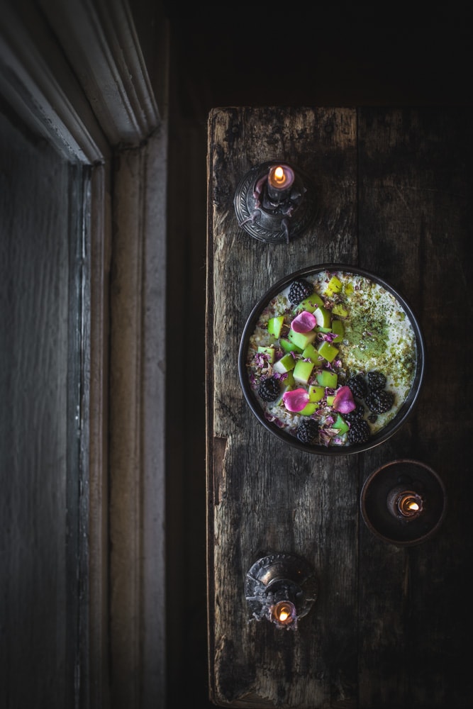 Bowl of cardamom rose porridge next to window with matcha dust, sprouted pumpkin seed butter, green apple, blackberries, and black candles